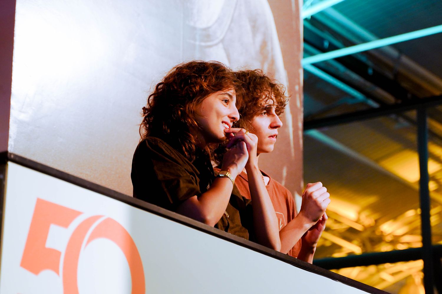 A young couple watches eagerly from a balcony at an indoor event.