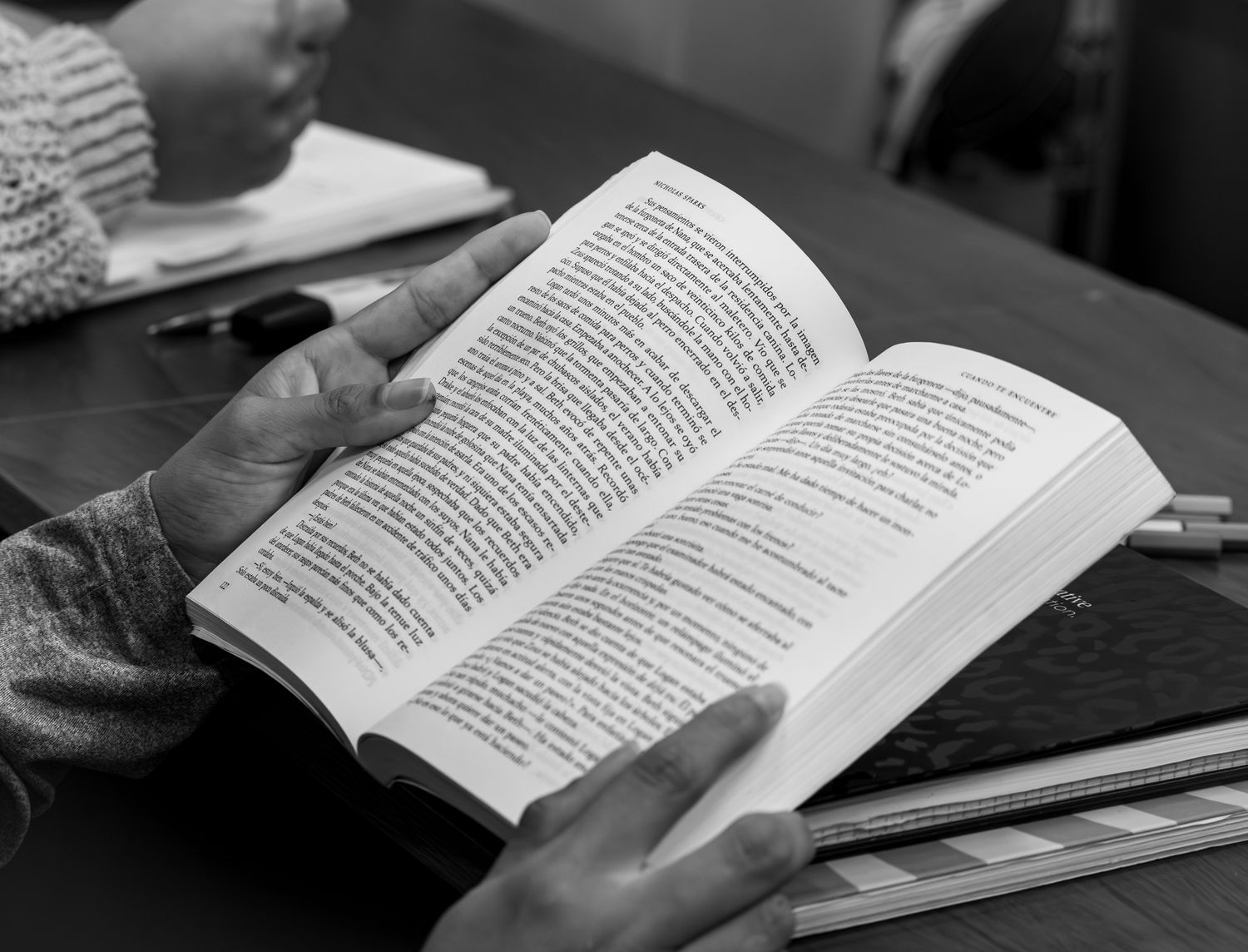 Monochrome photo of hands reading a book at a desk, evoking study and focus.