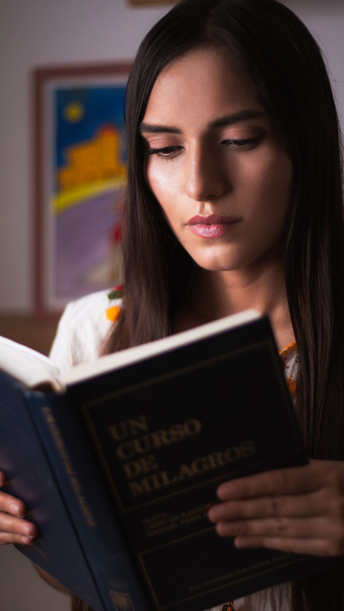 A young woman intently reading a book indoors in Cancún, Mexico, with a colorful artwork in the background.