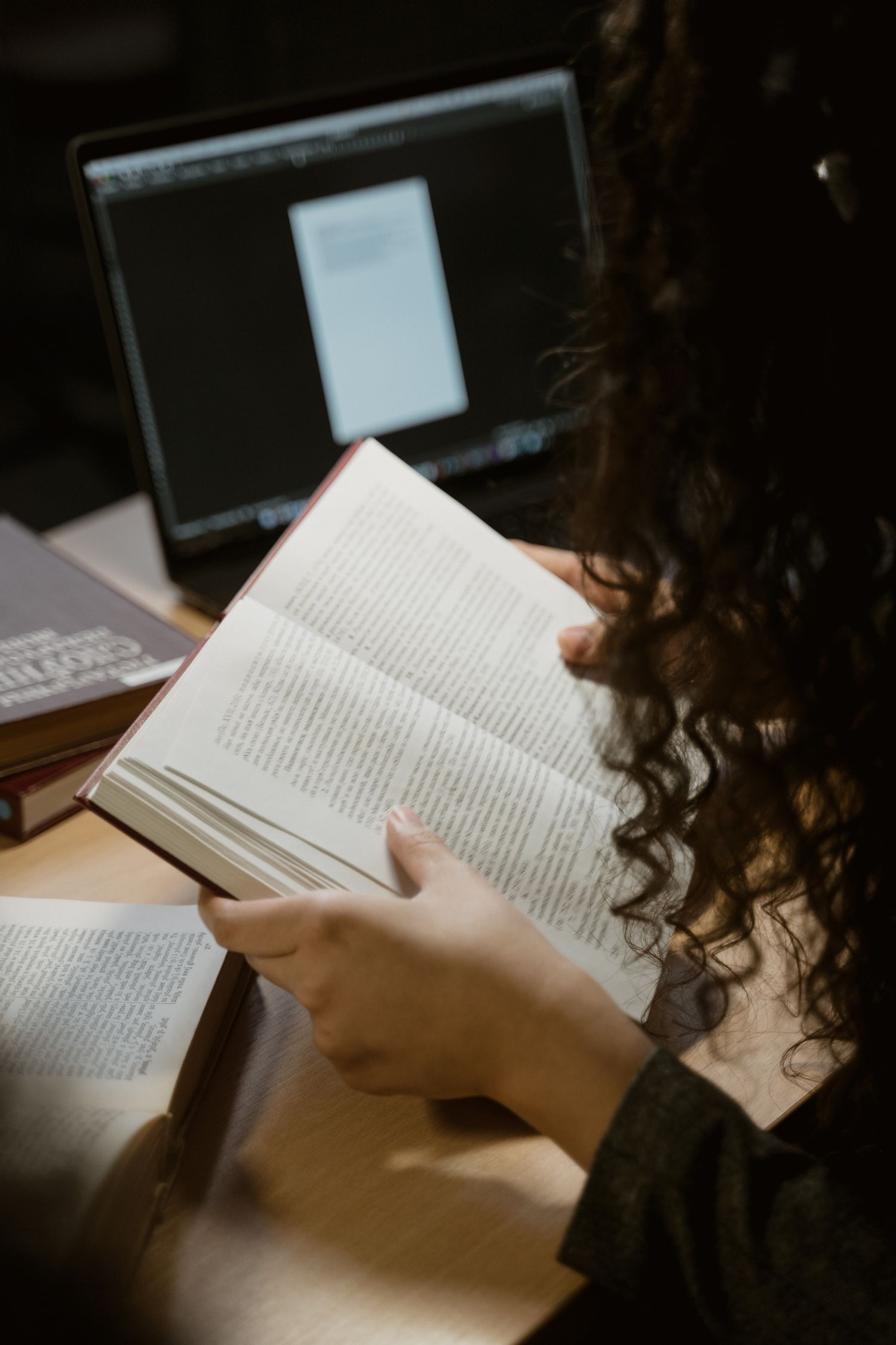 Focused woman reading a book at a wooden desk with books and laptop nearby.