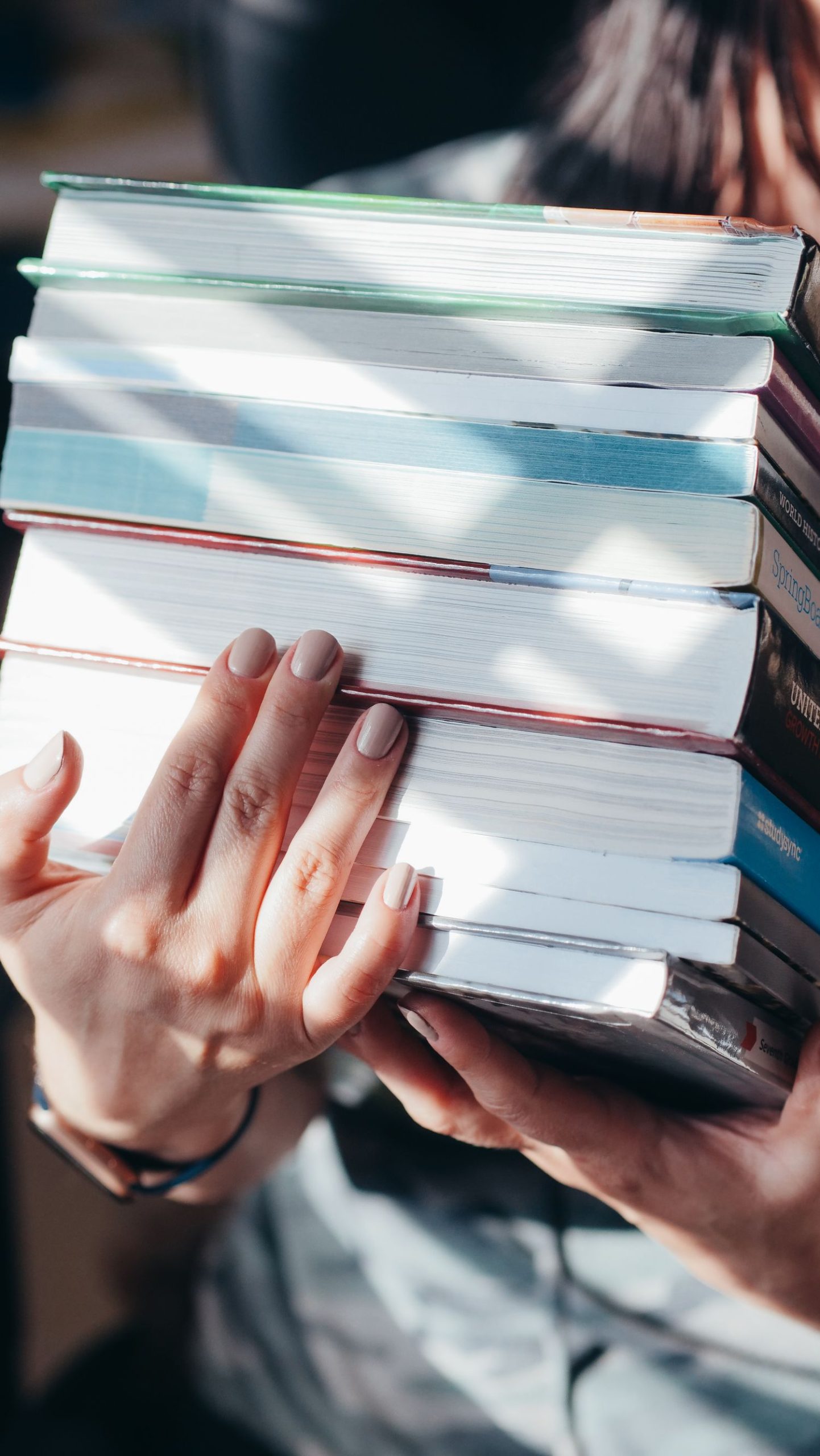 Close-up of hands holding a stack of diverse books under natural light indoors.